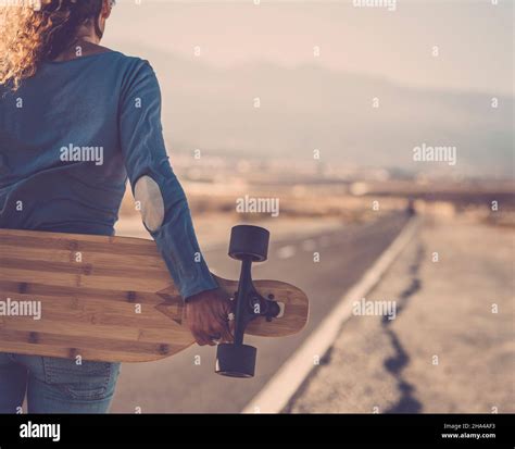 Carefree Woman Standing And Holding Wooden Longboard Behind Her Back On