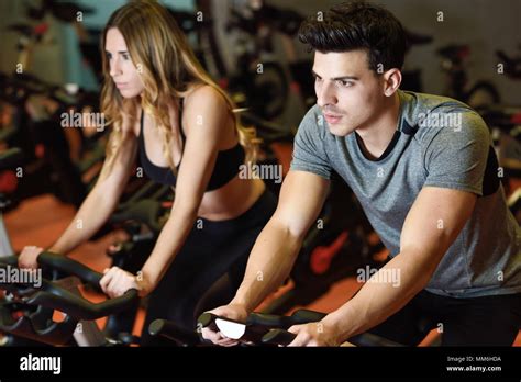 Young Man And Woman Biking In The Gym Exercising Legs Doing Cardio Workout Cycling Bikes Two