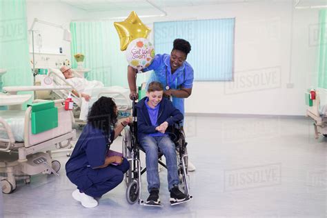 Doctor And Nurse Pushing Boy Patient In Wheelchair In Hospital Ward