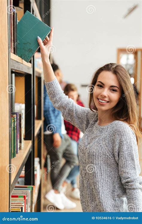 Young Smiling Woman Student Standing In Library Holding Book Stock