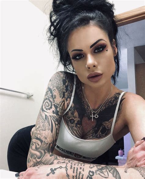A Woman With Tattoos On Her Arm And Chest Sitting In Front Of A Mirror Looking At The Camera