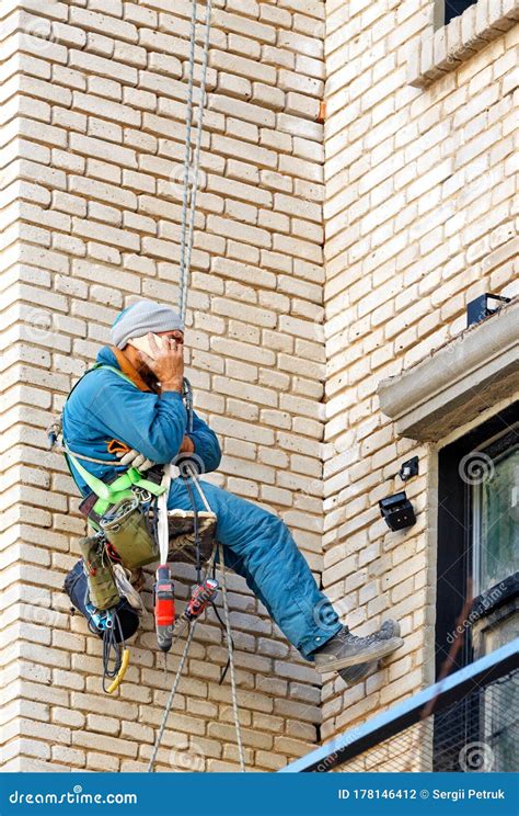 Builder Climber Hanging On Safety Ropes On The Wall Of An Old Brick Building With Tools Tied To