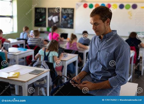 Male School Teacher Sitting On A Desk In An Elementary School Classroom