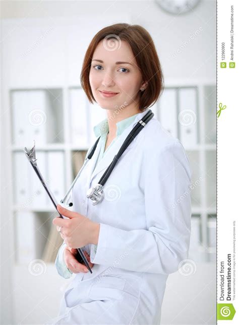 Young Brunette Female Doctor Standing With Clipboard And Smiling In