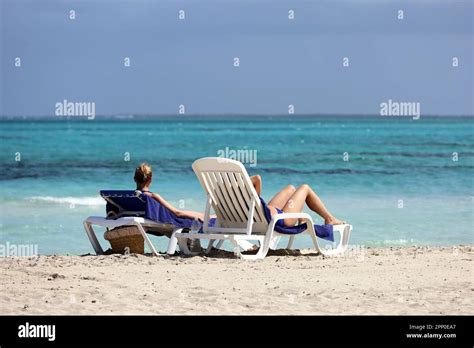 Two Girls In Bikini Tanning On White Deck Chairs On Sea Waves Background Vacation On Sandy