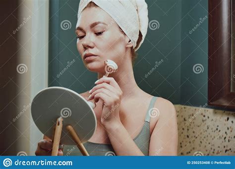 Young Woman Using Jade Facial Roller For Face Massage Sitting In Bathroom Looking In The Mirror