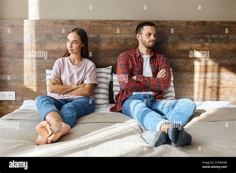 Young Frustrated Couple Sitting On Big Bed Keeping Hands And Legs Crossed Over Looking At