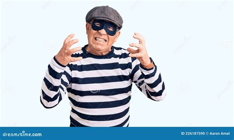 senior handsome man wearing burglar mask and t shirt shouting frustrated with rage hands trying