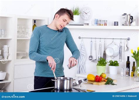 hombre joven que cocina una comida y que habla en el teléfono en la cocina imagen de archivo
