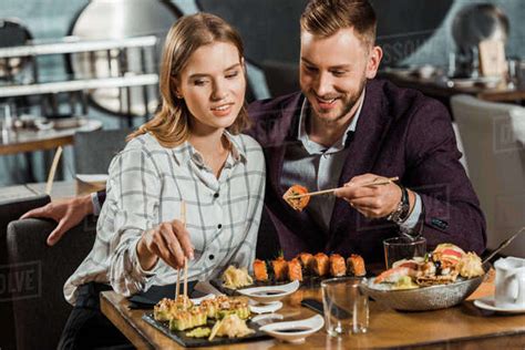 Happy Smiling Young Adult Couple Eating Sushi Rolls In Restaurant