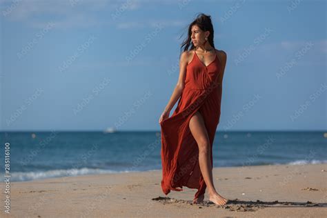 Pretty Young Woman In A Red Dress And Nude Foots On The Sand Of The