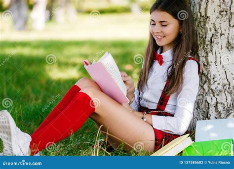 Beautiful European Schoolgirl Sits In A Park Near A Tree On The Green Grass And Reads A Book