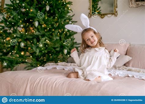 Pretty Cute Girl Is Sitting On Bed Next To Decorated Christmas Tree