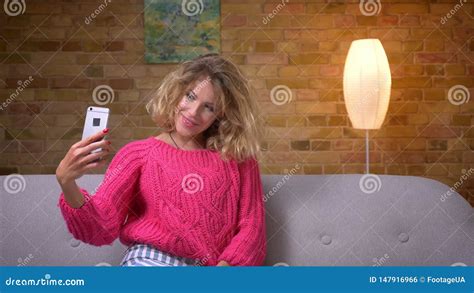blonde housewife in pink sweater making beautiful selfies on smartphone in cozy home atmosphere