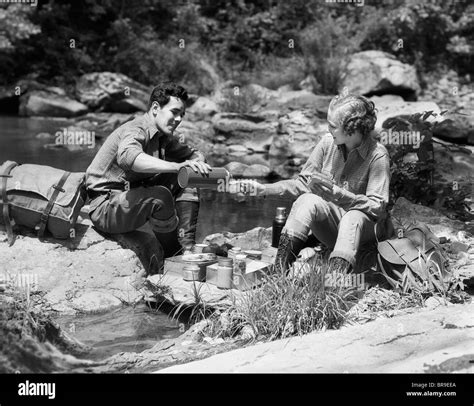 1930s Couple Having Picnic Sitting On Rocks By Stream Man Pouring Drink For Woman From Thermos