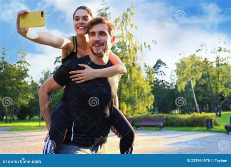 Happy Young Man Carrying His Girlfriend On His Back After Training In Park And She Taking Selfie