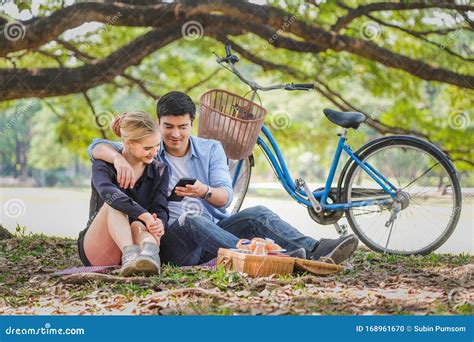 Young Couple In Love Sitting In Autumn Park Leaning Against A Tree Embracing One Another Stock