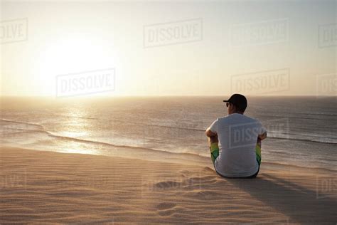 Rear View Of Man Sitting On Beach Against Sea During Sunset Stock
