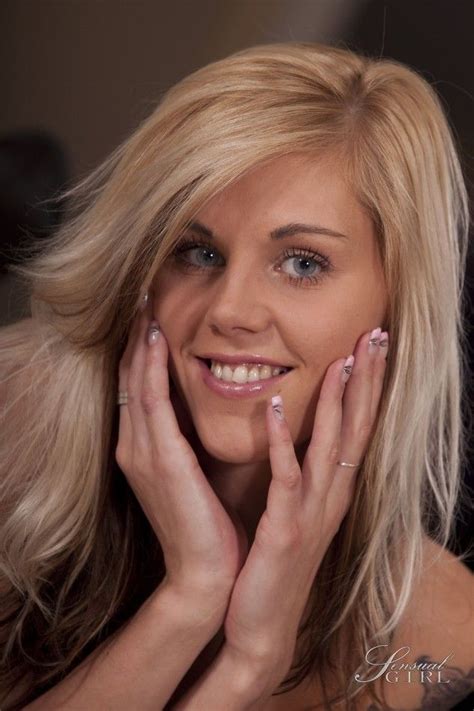 a woman with blonde hair and blue eyes smiles while holding her hands to her face