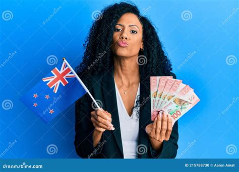 middle age african american woman holding new zealand flag and dollars looking at the camera