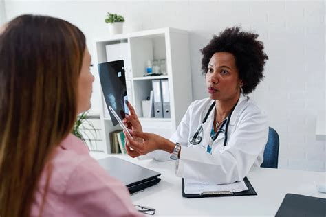 Doctor Talking With Patient At Desk In Medical Office Askmigration