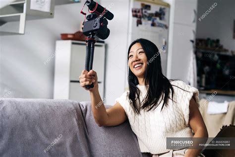 Smiling Ethnic Female Vlogger Recording Video On Photo Camera While Sitting On Couch In Living