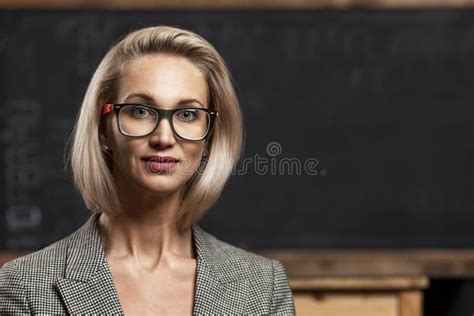 A Young Blonde Woman Teacher With Glasses Stands In A Class Near A Black School Board Distance