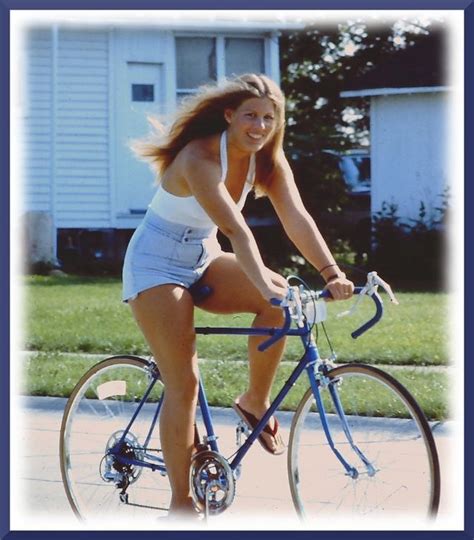 A Woman Riding A Bike Down A Street Next To A Grass Covered Field And