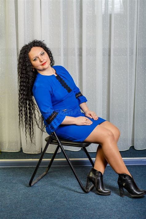 Curly Brunette Woman In Blue Sits On A Chair With Her Head Thrown Back And Stretched Her Legs