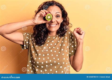 middle age beautiful woman holding kiwi over eye screaming proud celebrating victory and