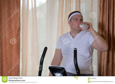 Fat Guy Exercising On Stationary Training Bicycle And Drinking Water