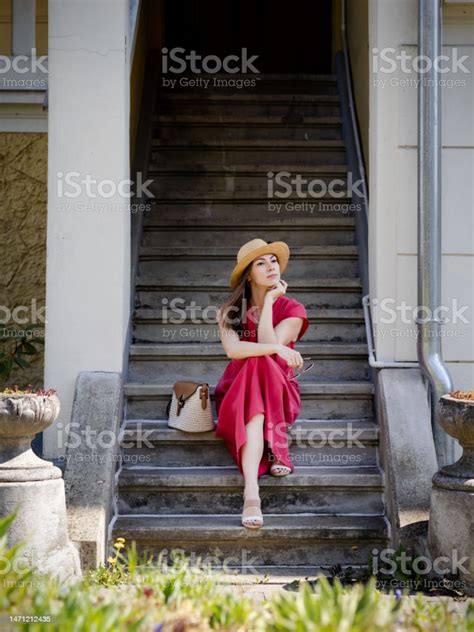 piękna młoda kobieta w eleganckiej czerwonej sukience i słomkowym kapeluszu siedzi na schodach