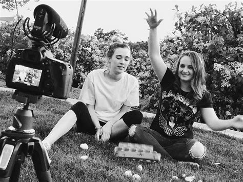 Two People Sitting On The Grass With Their Hands Up In Front Of Them And Cameras