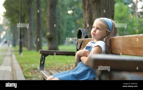 Small Irritated Child Girl Sitting Alone On A Bench In Summer Park