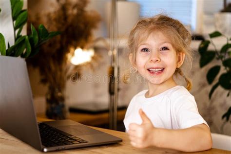A Small Blonde Girl With A Laptop Sits At A Table And Smiles Stock