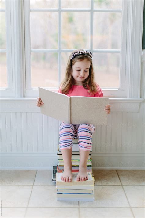 Cute Young Girl Sitting On A Stack Of Books Reading By Stocksy