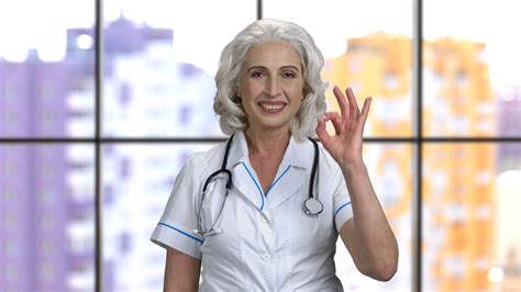 old senior female doctor wearing stethiscope and showing okay gesture windows background stock