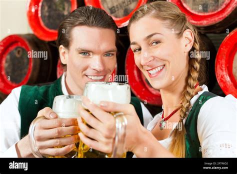 Young Couple Man And Woman In Traditional Bavarian Tracht Drinking Beer In A Brewery In Front