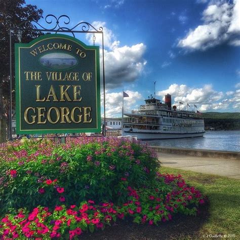 A Welcome Sign To The Village Of Lake George With A Boat In The Water