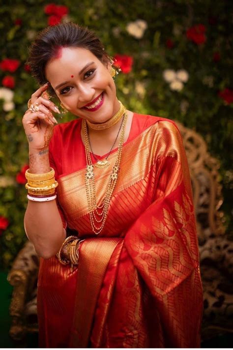 a woman in a red and gold sari smiles while standing next to a flower covered wall