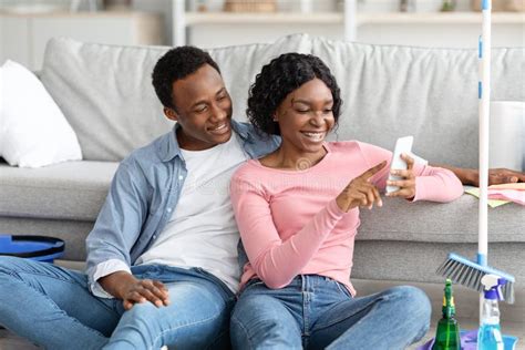 Black Lovers With Cleaning Tools Sitting On Floor Using Smartphone