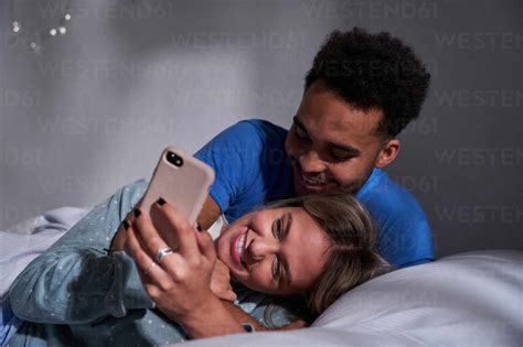delighted multiracial couple lying on bed while browsing smartphone and having fun together in