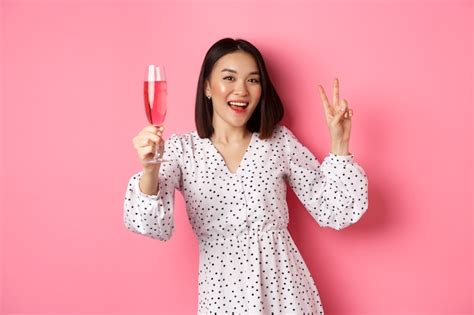 Праздник красивая азиатская женщина пьет шампанское и улыбается стоя в платье на розовом фоне