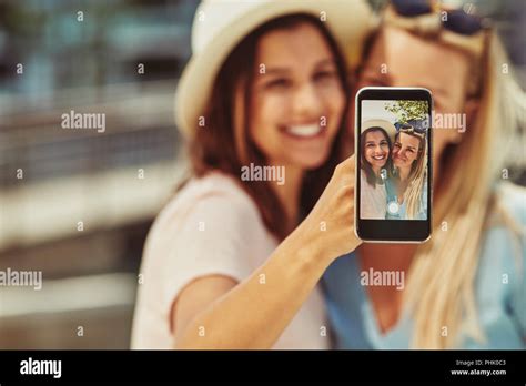 Two Smiling Young Female Friends Taking Selfies With A Cellphone While Enjoying A Day Out
