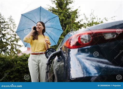 woman calling standing under umbrella while charging her electric car on the rainy day stock