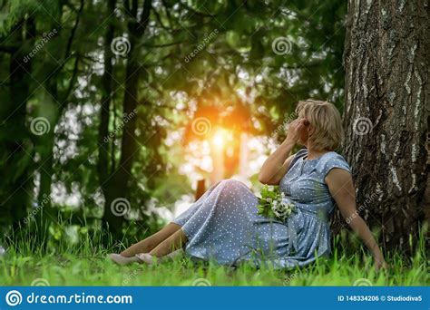 A Woman In A Blue Dress Sitting Near A Tree And Admires The Sunset