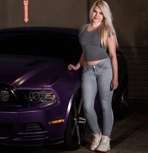 A Beautiful Blonde Woman Standing Next To A Purple Mustang In Front Of