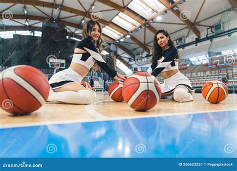 Low Angle Shot Of Two Cheerleaders Sitting With Basketballs On The Gymnasium Floor Sport