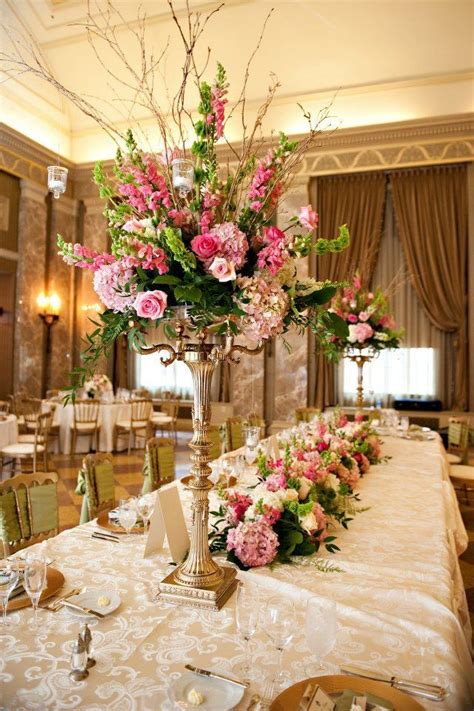 A Tall Vase Filled With Lots Of Pink Flowers On Top Of A Table Covered