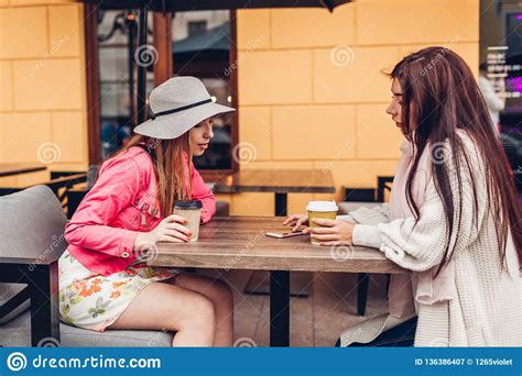 Two Women Chatting While Having Coffe In Outdoor Cafe. Happy Friends Using Phone Checking ...